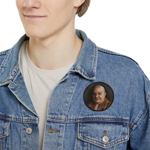 May include: A circular button badge displaying a portrait of a man in religious vestments. The badge has a black border and a detailed image of the man in a red and gold robe. The badge is pinned to a blue denim jacket.