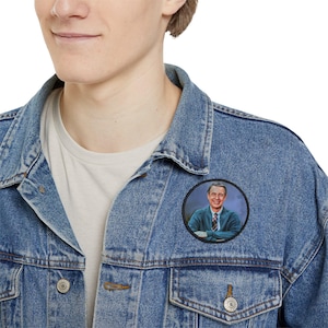 May include: A round patch featuring a portrait of a man in a blue sweater and a red tie. The patch is attached to a denim jacket. The portrait is set against a dark blue background within a black circular border.