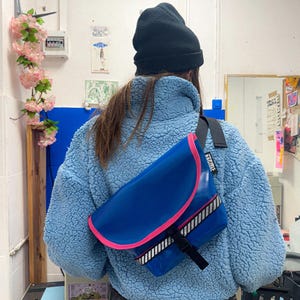 Peut inclure: Une personne portant une veste en polaire bleue et un bonnet noir porte un sac messager bleu, rose et blanc avec le mot "Timbuk2" imprimé sur la bandoulière.