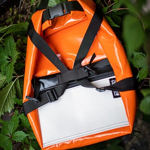 Puede incluir: Mochila naranja brillante con correas y hebillas negras. La bolsa tiene un panel blanco en la parte delantera y está rodeada de hojas verdes. La bolsa parece estar hecha de un material impermeable.