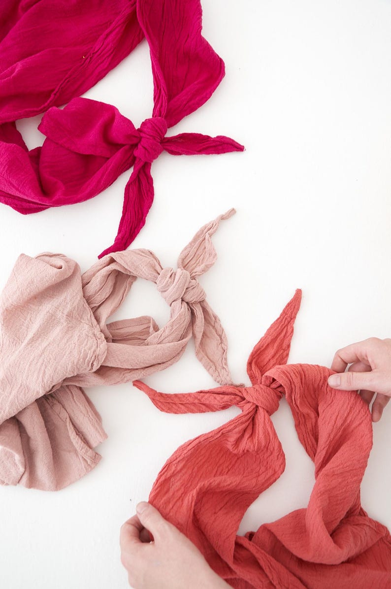 K&ouml;nnte beinhalten: Drei geknotete Stoffstirnb&auml;nder in Rosa- und Rott&ouml;nen werden auf einer wei&szlig;en Oberfl&auml;che pr&auml;sentiert. Die Stirnb&auml;nder sind aus einem strukturierten Stoff gefertigt, eines in tiefem Magenta, eines in hellem Rosa und eines in Koralle.