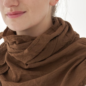 K&ouml;nnte beinhalten: Nahaufnahme eines braunen Schals, der um den Hals einer Person drapiert ist. Der Schal besteht aus einem strukturierten Stoff und hat eine warme, erdige braune Farbe. Der Schal ist in Schichten gewickelt, wodurch ein weicher, volumin&ouml;ser Look entsteht. Der Hintergrund ist schlicht wei&szlig;.