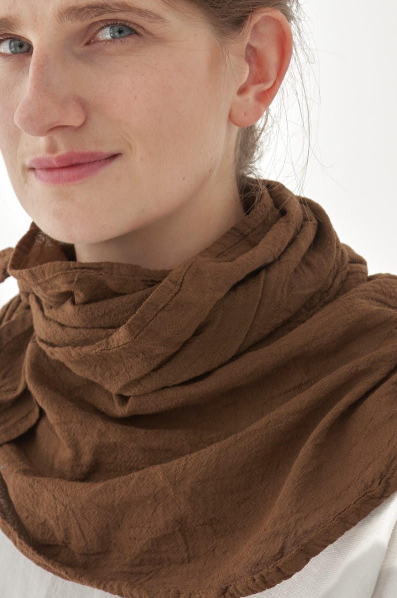 K&ouml;nnte beinhalten: Nahaufnahme eines braunen Schals, der um den Hals einer Person drapiert ist. Der Schal besteht aus einem strukturierten Stoff und hat eine warme, erdige braune Farbe. Der Schal ist in Schichten gewickelt, wodurch ein weicher, volumin&ouml;ser Look entsteht. Der Hintergrund ist schlicht wei&szlig;.