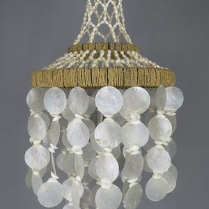 Può includere: Un mobile o lampada a sospensione con una struttura superiore conica avvolta in corda di juta color marrone chiaro e decorata con piccole conchiglie bianche. Sotto, molteplici fili di dischi circolari traslucidi in madreperla pendono a livelli.