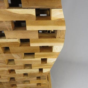Könnte beinhalten: Ein Beistelltisch aus zahlreichen kleinen, rechteckigen, hellbraunen Holzblöcken, die in einem ineinandergreifenden Gittermuster gestapelt sind. Die Struktur weist eine geschwungene, sanduhrartige Silhouette mit einer flachen Oberfläche auf.