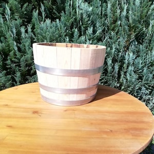 Könnte beinhalten: Ein hellfarbener, fassförmiger Holzpflanzkübel mit zwei silbernen Metallbändern in der Mitte. Der Pflanzkübel steht auf einem runden Holztisch. Der Hintergrund ist eine dichte grüne Hecke.