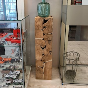 Könnte beinhalten: Ein hoher, rechteckiger Holzsockel mit natürlicher, hellbrauner Oberfläche. Der Sockel besteht aus unregelmäßig geformten Holzblöcken. Auf dem Sockel steht eine große, grüne Glasflasche. Im Hintergrund eine Glasvitrine mit Brillen.