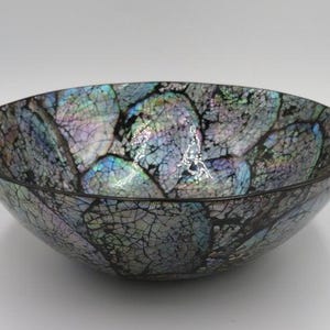 Op de afbeelding: Een ronde kom met een decoratief mozaïekoppervlak bestaande uit iriserende schelpfragmenten. De buiten- en binnenkant vertonen een gebarsten patroon met glinsterende tinten blauw, paars en groen, omlijnd door donkere, dunne voeglijnen.