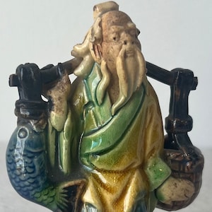Op de afbeelding: Een keramieken beeldje van een oude man die een vis en twee manden op een draagstok draagt. De figuur draagt een groene en gele jurk met een blauwe broek. De vis is blauw en groen. Het beeldje staat op een witte ondergrond.