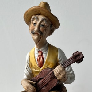 May include: A ceramic figurine of an elderly man playing a small guitar. He wears a brown hat, yellow vest, white shirt, and brown pants. The figurine is on a decorative base with a musical theme.