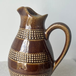 May include: A brown ceramic pitcher with a handle and spout. The pitcher has a glossy finish and is decorated with horizontal bands of small, raised dots. The overall design is a warm, earthy tone, suitable for serving beverages.