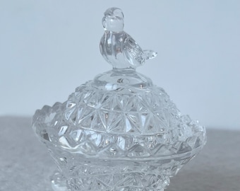 May include: A clear, cut-glass trinket dish with a bird-shaped lid finial. The dish has a diamond-cut pattern and sits on three small feet. The lid is domed, and the bird is perched atop. A decorative piece.