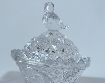 May include: A clear pressed glass candy dish featuring a faceted diamond pattern and a scalloped rim. The lid is topped with a small glass bird figurine, and the entire piece rests on a circular pedestal base with a starburst design.