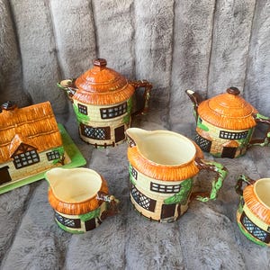 May include: A vintage ceramic tea set featuring a cottage design. The set includes a teapot, creamer, sugar bowl, and two cups. The pieces are painted with brown, orange, and green details, resembling a thatched roof and brickwork.