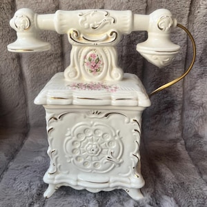 Puede incluir: Un teléfono de cerámica blanco roto de estilo vintage con detalles dorados. El teléfono presenta un diseño floral y detalles ornamentados. El auricular está conectado a la base con un cable metálico dorado. Objeto decorativo.