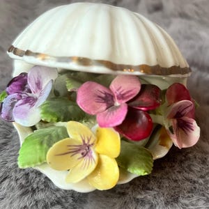 May include: A white porcelain shell-shaped trinket box with a gold trim. The box is filled with colourful, detailed porcelain pansies in shades of purple, pink, and yellow, with green leaves. The box is open, revealing the flowers.