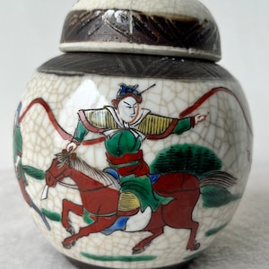 May include: A small, round ceramic jar with a lid. The jar is off-white with a crackle glaze and features a painted scene of a warrior on horseback. The warrior wears green and red clothing. The lid is brown and white.