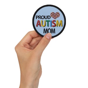 May include: A round patch with the words "PROUD AUTISM MOM" in black, green, red, blue, and yellow. A heart made of colorful puzzle pieces is above the word "AUTISM". The patch has a light blue background and a black border.