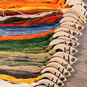 May include: A collection of embroidery floss skeins in a spectrum of colors including red, orange, yellow, green, blue, brown, and white. Each skein is organized on a light-colored wooden organizer shaped like a teardrop with a circular hole.
