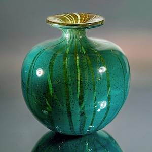 Op de afbeelding: Een turquoise glazen vaas met een ronde body en een smalle hals. Verticale groene strepen lopen van de hals naar de basis. De rand is lichtbruin van kleur. De vaas staat op een reflecterend oppervlak.