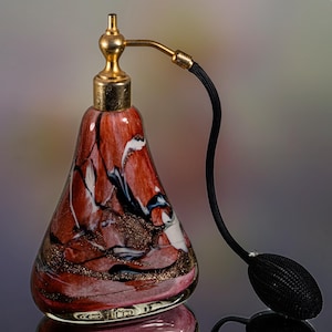 Op de afbeelding: Een decoratieve parfumfles met een rood, wit en zwart gemarmerd ontwerp. De fles heeft een goudkleurige metalen bovenkant en een zwarte verstuiver. De basis van de fles is lichtroze met gouden accenten.