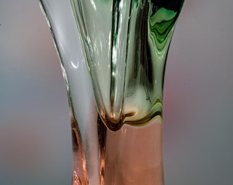 May include: A tall, elegant glass vase with a unique, organic shape. The vase transitions from a clear top to a green and then a peach-colored base. The design features flowing lines and a flared opening, creating a modern aesthetic.
