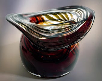 May include: A decorative glass vase with a unique design. The vase features a deep burgundy base with swirling patterns and a silver-rimmed top. The top has a wave-like shape with gold and silver accents, creating a striking visual contrast.