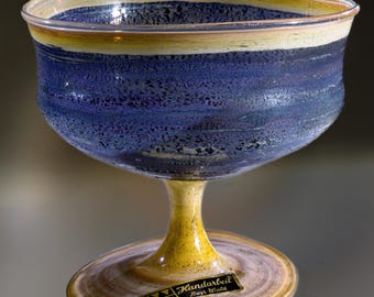 May include: A handcrafted glass goblet with a wide bowl and a slender stem. The bowl features horizontal bands of blue, yellow, and clear glass. The stem and base are yellow. A small label with text is affixed to the base.