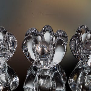 Könnte beinhalten: Drei klare, blumenförmige Kerzenhalter aus Glas. Jeder Halter hat einen abgerundeten Sockel und eine Oberseite mit sechs blütenblattartigen Abschnitten. Das Glas reflektiert das Licht und erzeugt einen schimmernden Effekt. Die Kerzenhalter werden auf einer reflektierenden Oberfläche ausgestellt.