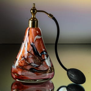 Könnte beinhalten: Eine Vintage-Parfümflasche aus Glas mit einem goldenen Zerstäuber und einer schwarzen Sprühdüse. Die Flasche ist birnenförmig und hat ein wirbelndes Muster in Rosa, Weiß und Schwarz.