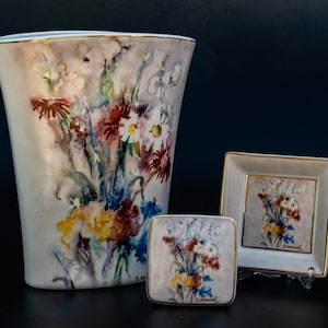 Op de afbeelding: Een set van drie porseleinen vazen met een aquarel bloemenontwerp. De grootste vaas is hoog en slank, terwijl de twee kleinere vazen vierkant zijn. Alle drie vazen tonen een boeket kleurrijke bloemen, waaronder rode, gele, blauwe en witte.