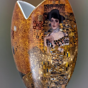 Könnte beinhalten: Eine ovale Keramikvase mit einem Porträt einer Frau mit schwarzem Hut, mit goldenen und braunen Akzenten. Die Vase hat eine einzigartige Form und ein glänzendes Finish, was sie zu einem dekorativen Stück macht.