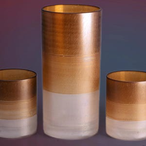 Könnte beinhalten: Ein Set aus fünf dekorativen Glaszylindern. Die Zylinder haben einen metallisch goldenen oberen Teil, einen Farbverlauf aus Gold und Beige in der Mitte und eine weiße Basis. Der höchste Zylinder befindet sich in der Mitte, mit kürzeren Zylindern auf beiden Seiten.