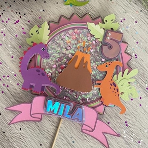 Könnte beinhalten: Ein rosa-grüner Dinosaurier-Kuchentopper mit einem Vulkan, zwei Dinosauriern und dem Namen "Mila" auf einem rosa Banner. Der Topper ist mit Glitzer und Pailletten verziert.