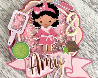 May include: A pink and gold cake topper featuring a cartoon girl with brown skin and black hair wearing a pink robe. The topper also includes a makeup brush, a cucumber slice, a spa stone, and a banner that says "Amy".