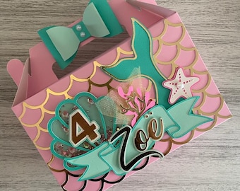 May include: Pink favor box with a mermaid tail, seashells, and a starfish. The box is decorated with gold glitter and the number 4. The name Zoe is written on the box in black.