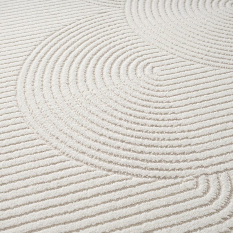 May include: Cream-coloured rug featuring a repeating pattern of curved lines. The lines create a textured surface with a soft, plush appearance. The design is a series of concentric arcs and straight lines, giving a modern, minimalist aesthetic.