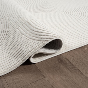 May include: A rolled-up, cream-coloured rug with a textured, circular pattern. The rug's design features concentric circles and parallel lines, creating a tactile and visually appealing surface. The rug is displayed on a dark wood floor.