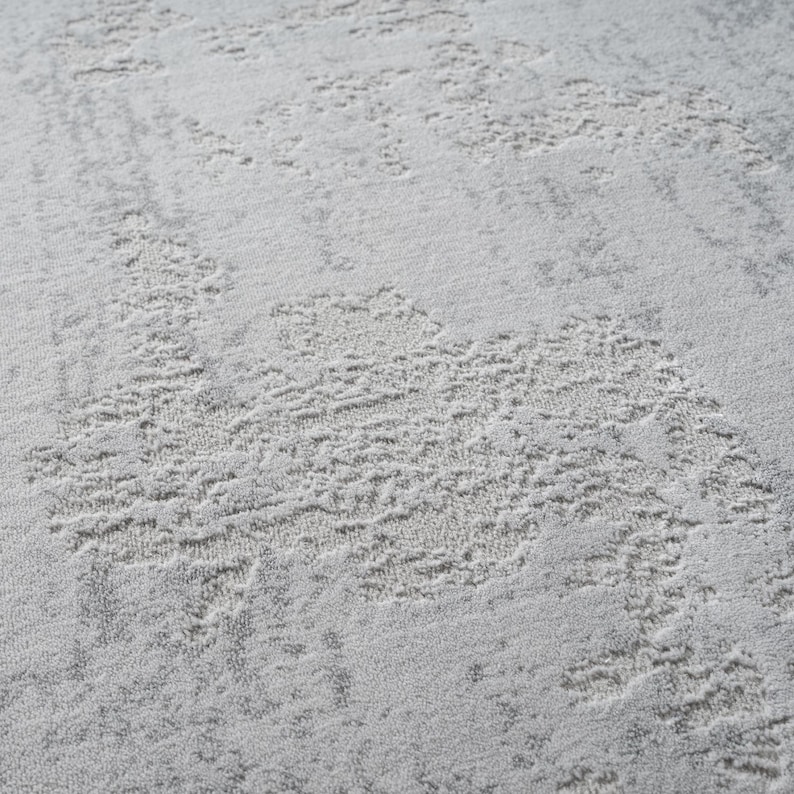 May include: Close-up of a light gray rug with a textured, abstract design. The pattern features raised, irregular shapes in a lighter shade of gray, creating a subtle, layered effect. The rug's surface appears soft and plush, suitable for home decor.