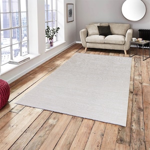 Könnte beinhalten: Ein weißer Teppich mit leichter Struktur, der auf einem Holzboden in einem Wohnzimmer liegt. Der Teppich liegt vor einem beigen Sofa mit einem Kissen und einem kleinen Beistelltisch.