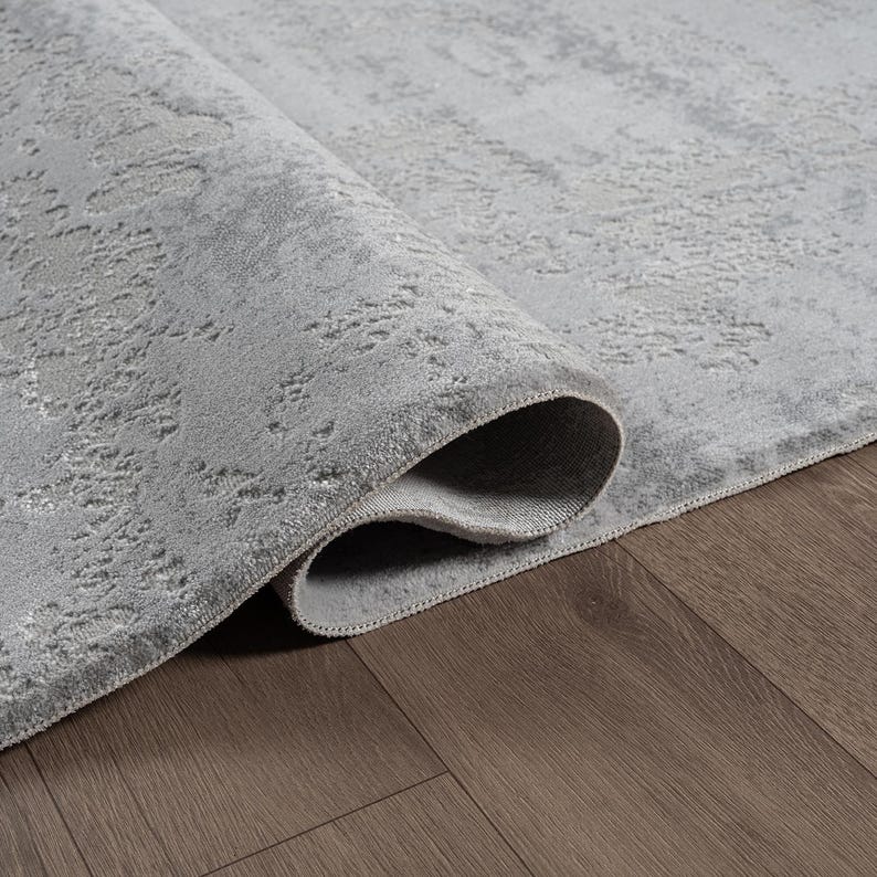 May include: A rolled-up, light gray area rug with a textured, distressed pattern. The rug's edge is visible, revealing a darker gray backing. The rug is placed on a dark brown wooden floor.