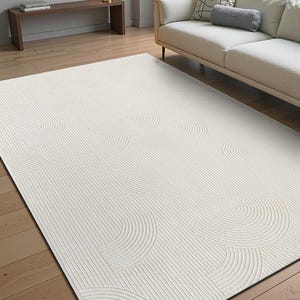 May include: Cream-coloured area rug with a textured design. The rug features a pattern of curved lines and semi-circles, creating a layered effect. The rug is rectangular and sits on a wooden floor.