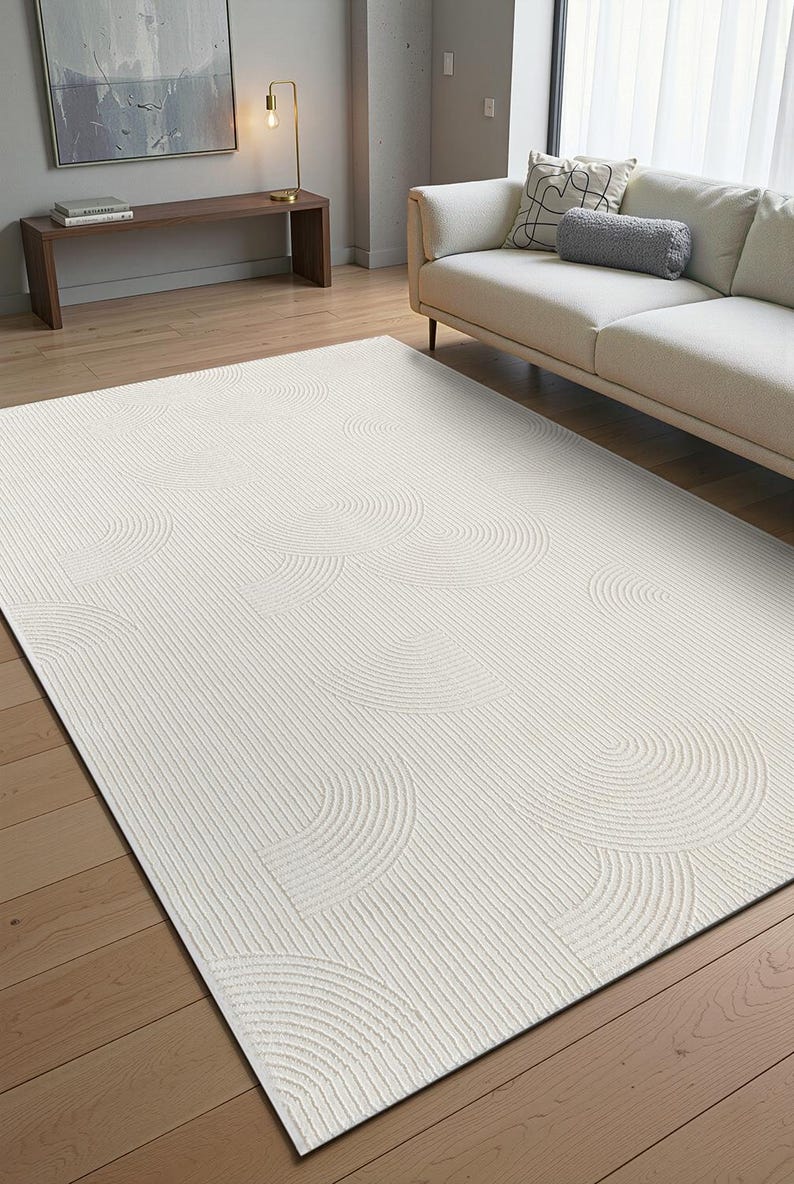 May include: Cream-coloured area rug with a textured design. The rug features a pattern of curved lines and semi-circles, creating a layered effect. The rug is rectangular and sits on a wooden floor.