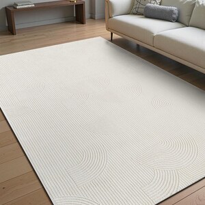 May include: Cream-coloured area rug with a textured design. The rug features a pattern of curved lines and semi-circles, creating a layered effect. The rug is placed on a wooden floor, in a living room setting.