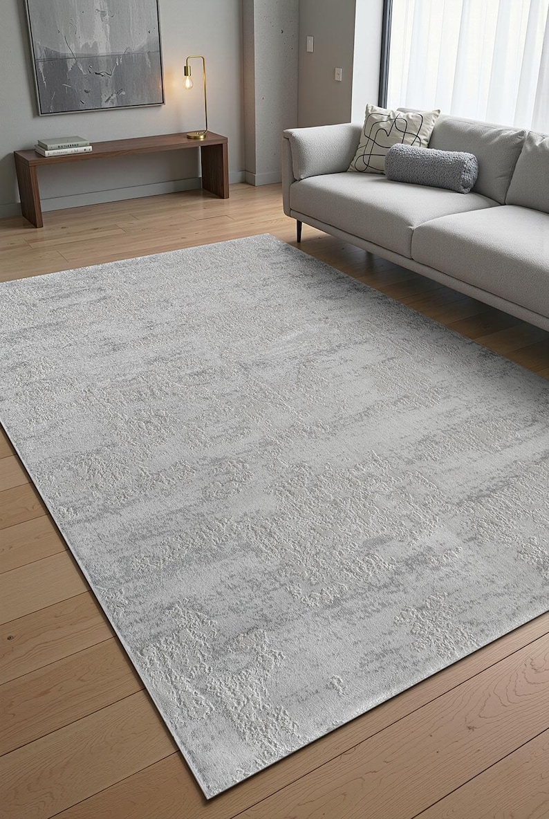 May include: A large, rectangular area rug with a textured, light gray design. The rug is placed on a hardwood floor, and the overall aesthetic is modern and minimalist. The rug's dimensions are not visible.