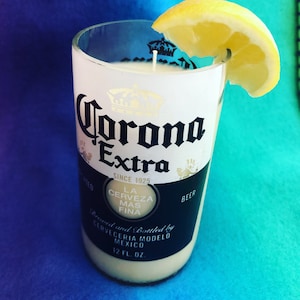 May include: A white candle in a clear glass bottle with a Corona Extra beer label. The bottle is decorated with a gold crown and the text "La Cerveza Mas Fina". A slice of lemon is placed on top of the candle.