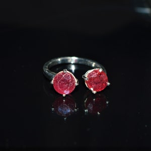 May include: Silver open ring with two large, round, red gemstones. The gemstones are held in place by prongs, and the ring has a textured band. The ring is shown on a reflective black surface.