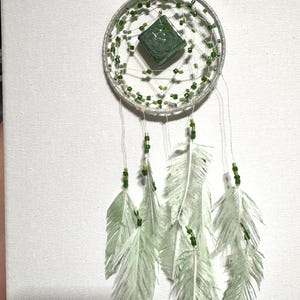 May include: A green and white dreamcatcher with feathers hanging from the bottom. The dreamcatcher has a green glass bead in the center.