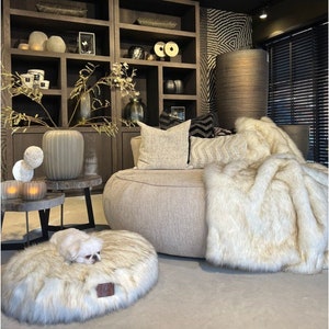 May include: A light brown, round, faux fur dog bed with a white dog lying on it. The bed is in a living room with a large, round, light brown sofa with a white faux fur throw blanket on it. There are two dark brown wooden shelves with various decorative items on them.