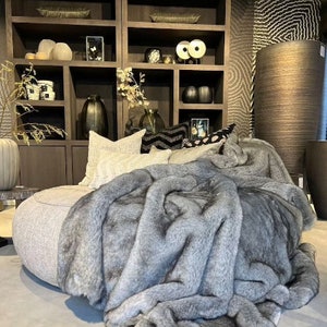 Stylish interior in subtle gray tones featuring luxurious home decor elements that exude comfort and design.  Ash-colored furry throw accentuates the refined ambiance, while a comfortable sofa surrounded by designer cushions and modern accents creates an inviting, cozy space. Minimalist decor paired with exclusive accent pieces and a carefully curated arrangement ensures every detail radiates sophisticated elegance, perfect for those who appreciate refined comfort and innovative design.